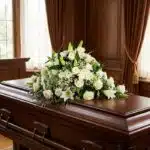 Cubre caja fúnebre solemne con rosas blancas, lirios y follaje verde sobre un ataúd de madera oscura en un entorno elegante y