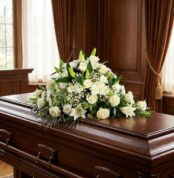 Cubre caja fúnebre solemne con rosas blancas, lirios y follaje verde sobre un ataúd de madera oscura en un entorno elegante y