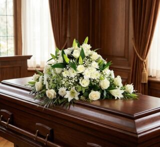 Cubre caja fúnebre solemne con rosas blancas, lirios y follaje verde sobre un ataúd de madera oscura en un entorno elegante y