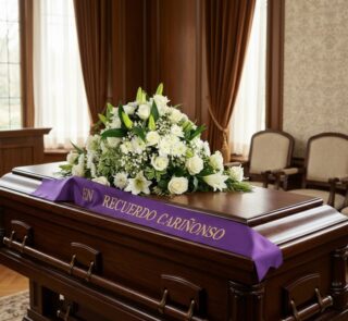 Cubre caja fúnebre solemne con arreglo floral blanco y cinta morada 'En Recuerdo Cariñoso' en sala de velación premium.