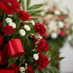 Primer plano de arreglo fúnebre con rosas rojas, gerberas y crisantemos blancos, con rocío sutil y cinta satinada roja. Fondo