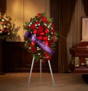 Pedestal fúnebre con rosas rojas, margaritas blancas y follaje verde, adornado con una cinta morada que dice 'Con Sentido Pés