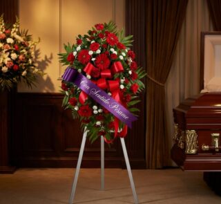 Pedestal fúnebre con rosas rojas, margaritas blancas y follaje verde, adornado con una cinta morada que dice 'Con Sentido Pés