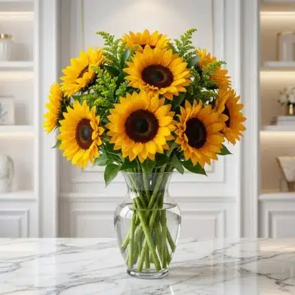 Jarrón de cristal transparente con doce girasoles grandes y vibrantes, hojas verdes y follaje decorativo, sobre una mesa de m