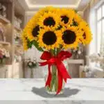 Jarrón de cristal con doce girasoles grandes y vibrantes, atado con un lazo rojo, sobre una mesa de mármol en una floristería