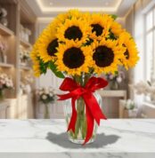 Jarrón de cristal con doce girasoles grandes y vibrantes, atado con un lazo rojo, sobre una mesa de mármol en una floristería