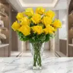 Jarrón de cristal tallado con doce rosas amarillas abiertas y follaje verde, sobre una superficie de mármol blanco en un ento