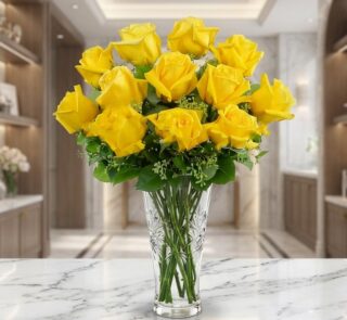 Jarrón de cristal tallado con doce rosas amarillas abiertas y follaje verde, sobre una superficie de mármol blanco en un ento