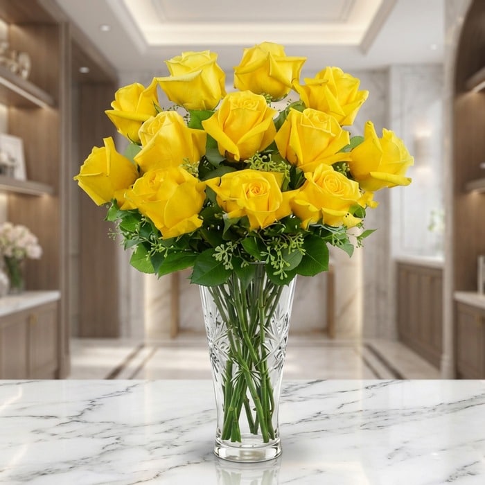 Jarrón de cristal tallado con doce rosas amarillas abiertas y follaje verde, sobre una superficie de mármol blanco en un ento