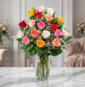 Jarrón de cristal con 18 rosas de diversos colores (rojo, rosa, amarillo, blanco, naranja) y follaje verde, sobre una mesa de