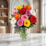 Jarrón de cristal transparente con un vibrante arreglo de gerberas en tonos rojos, naranjas, amarillos, rosas y blancos, sobr