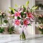 Jarrón de cristal con un exuberante arreglo de lirios rosas y blancos, follaje verde, y pequeñas flores moradas y blancas, at