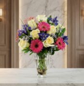 Jarrón Primavera con un vibrante arreglo floral de rosas blancas, gerberas fucsias y lirios morados, sobre una superficie de