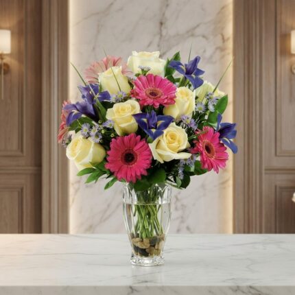 Jarrón Primavera con un vibrante arreglo floral de rosas blancas, gerberas fucsias y lirios morados, sobre una superficie de