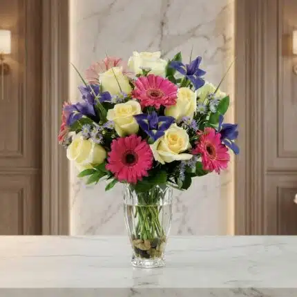 Jarrón Primavera con un vibrante arreglo floral de rosas blancas, gerberas fucsias y lirios morados, sobre una superficie de