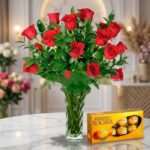 Jarrón de cristal con un exuberante arreglo de rosas rojas frescas y follaje verde, acompañado de una caja de bombones Ferrer