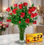 Jarrón de cristal con un exuberante arreglo de rosas rojas frescas y follaje verde, acompañado de una caja de bombones Ferrer