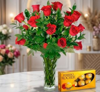 Jarrón de cristal con un exuberante arreglo de rosas rojas frescas y follaje verde, acompañado de una caja de bombones Ferrer