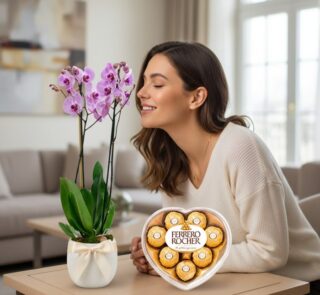 Mujer sonriendo y oliendo una orquídea GALA morada en maceta blanca con lazo, junto a una caja de chocolates Ferrero Rocher e