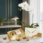 Arreglo MARELY con orquídea Phalaenopsis blanca, caja de chocolates Ferrero Rocher, tarjeta de regalo y cinta dorada sobre má