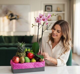 Mujer sonriente admirando el Arreglo ANTONELLO con orquídea Phalaenopsis rosa y frutas frescas como piña, manzanas y kiwis, e