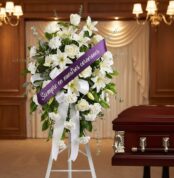 Pedestal conmemorativo de flores blancas, lirios y rosas, con cinta morada que dice 'Siempre en nuestros corazones', junto a