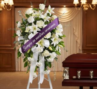 Pedestal conmemorativo de flores blancas, lirios y rosas, con cinta morada que dice 'Siempre en nuestros corazones', junto a