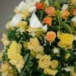 Fotografía macro premium del Pedestal Fúnebre Descansa Ahora, mostrando rosas amarillas y naranjas con rocío sutil, follaje v