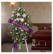 Pedestal fúnebre Esteban con rosas moradas y blancas, lirios, y cinta 'En Recuerdo Eterno' junto a un ataúd en sala de velaci