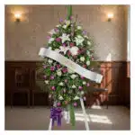 Pedestal fúnebre Esteban con arreglo floral de lirios blancos, rosas moradas y follaje verde, banda blanca con nombre 'Esteba