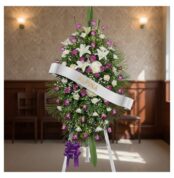 Pedestal fúnebre Esteban con arreglo floral de lirios blancos, rosas moradas y follaje verde, banda blanca con nombre 'Esteba