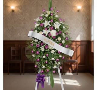 Pedestal fúnebre Esteban con arreglo floral de lirios blancos, rosas moradas y follaje verde, banda blanca con nombre 'Esteba