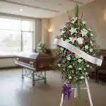 Pedestal fúnebre Esteban con rosas moradas y blancas, lirios y follaje, en una sala de velación moderna con ataúd desenfocado
