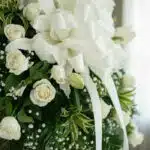 Primer plano macro de un arreglo fúnebre con rosas blancas, lirios y follaje verde con rocío, destacando texturas y una cinta