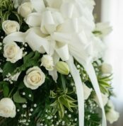 Primer plano macro de un arreglo fúnebre con rosas blancas, lirios y follaje verde con rocío, destacando texturas y una cinta