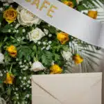 Arreglo floral fúnebre con rosas blancas y amarillas, follaje verde, rocío sutil y cinta satinada. Detalle de sobre de condol