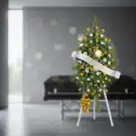 Pedestal fúnebre con rosas amarillas y blancas, follaje verde, y cinta blanca con el nombre 'COLCAFE', en una sala de velació