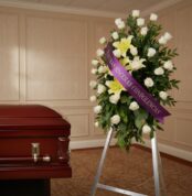 Pedestal fúnebre "Luz de Jacinta" con rosas blancas y lirios amarillos, cinta morada con mensaje de condolencia, junto a un a