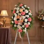 Pedestal fúnebre de homenaje para Timoteo, con arreglo floral de claveles coral, rosas durazno y crisantemos blancos, en sala