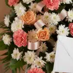 Pedestal fúnebre con rosas durazno, claveles coral y crisantemos blancos, adornado con cinta satinada. Un sobre de condolenci