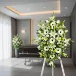 Pedestal fúnebre Inmaculada con lirios blancos, gerberas y follaje verde en una sala de velación moderna, transmitiendo paz y
