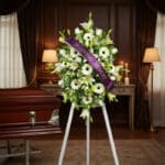 Pedestal fúnebre Inmaculada con lirios blancos, gerberas y follaje, adornado con una cinta morada que dice 'In Loving Memory'