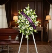 Pedestal fúnebre Inmaculada con lirios blancos, gerberas y follaje, adornado con una cinta morada que dice 'In Loving Memory'