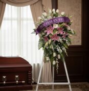 Pedestal fúnebre "Legado Eterno" con lirios rosados, rosas blancas y follaje, adornado con una cinta morada que dice "Descans