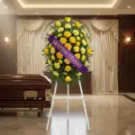 Pedestal fúnebre 'Legado Sereno' con rosas y claveles amarillos, cinta morada 'Sentido Pésame', junto a un ataúd en sala de v