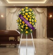 Pedestal fúnebre 'Legado Sereno' con rosas y claveles amarillos, cinta morada 'Sentido Pésame', junto a un ataúd en sala de v