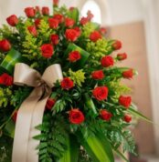 Primer plano de un pedestal fúnebre con rosas rojas vibrantes y follaje verde exuberante, adornado con una cinta satinada col