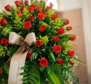 Primer plano de un pedestal fúnebre con rosas rojas vibrantes y follaje verde exuberante, adornado con una cinta satinada col