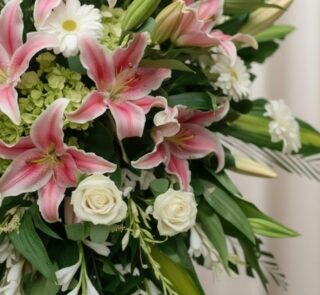 Primer plano macro de un arreglo fúnebre con lirios rosados, rosas blancas y margaritas, con rocío sutil y cinta satinada. Pr