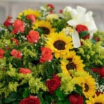 Fotografía macro premium del Pedestal Fúnebre Solemne, mostrando rosas rojas, girasoles amarillos y follaje verde con rocío s